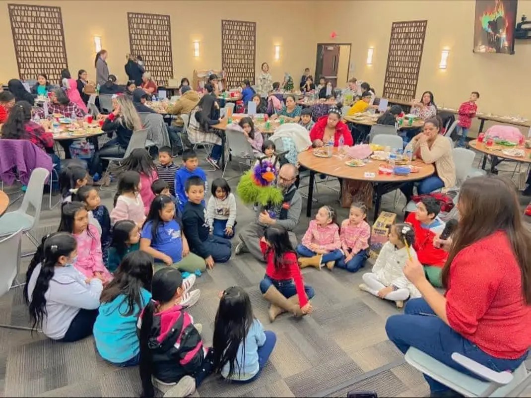 Photo from Puertas Abiertas showing a room full of mostly Latinx people. The front of the image shows a story time with two adults and many children playing with a puppet.