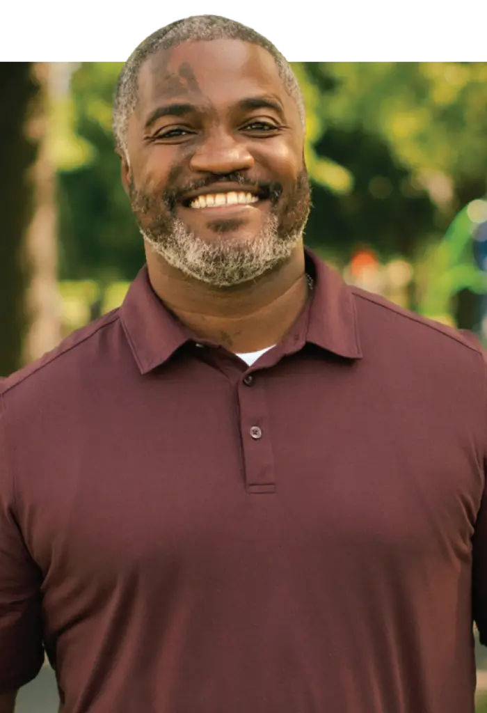 Smiling man with gray beard and short gray hair wearing a maroon polo shirt outdoors among blurred green trees