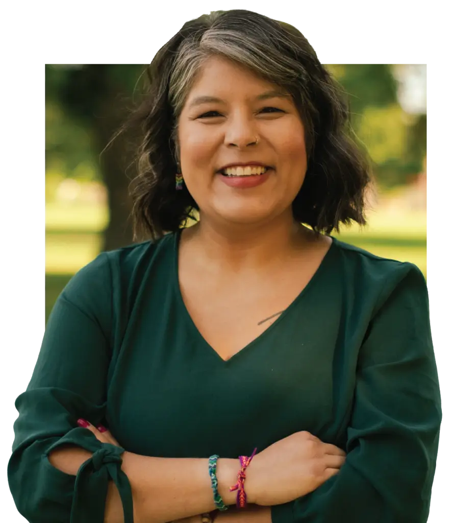 Smiling woman with shoulder-length wavy dark hair and gray streaks, wearing a dark green top and colorful woven bracelets, arms crossed outdoors.